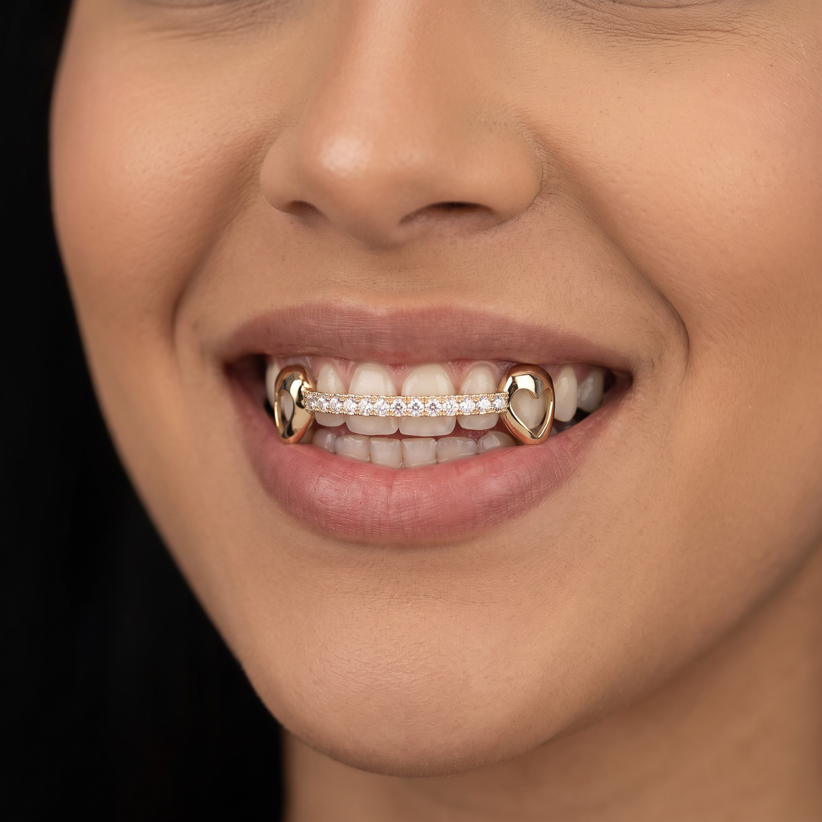 Smiling woman wearing a 6 piece yellow gold grill with heart cutout canines and a moissanite center, on a black background