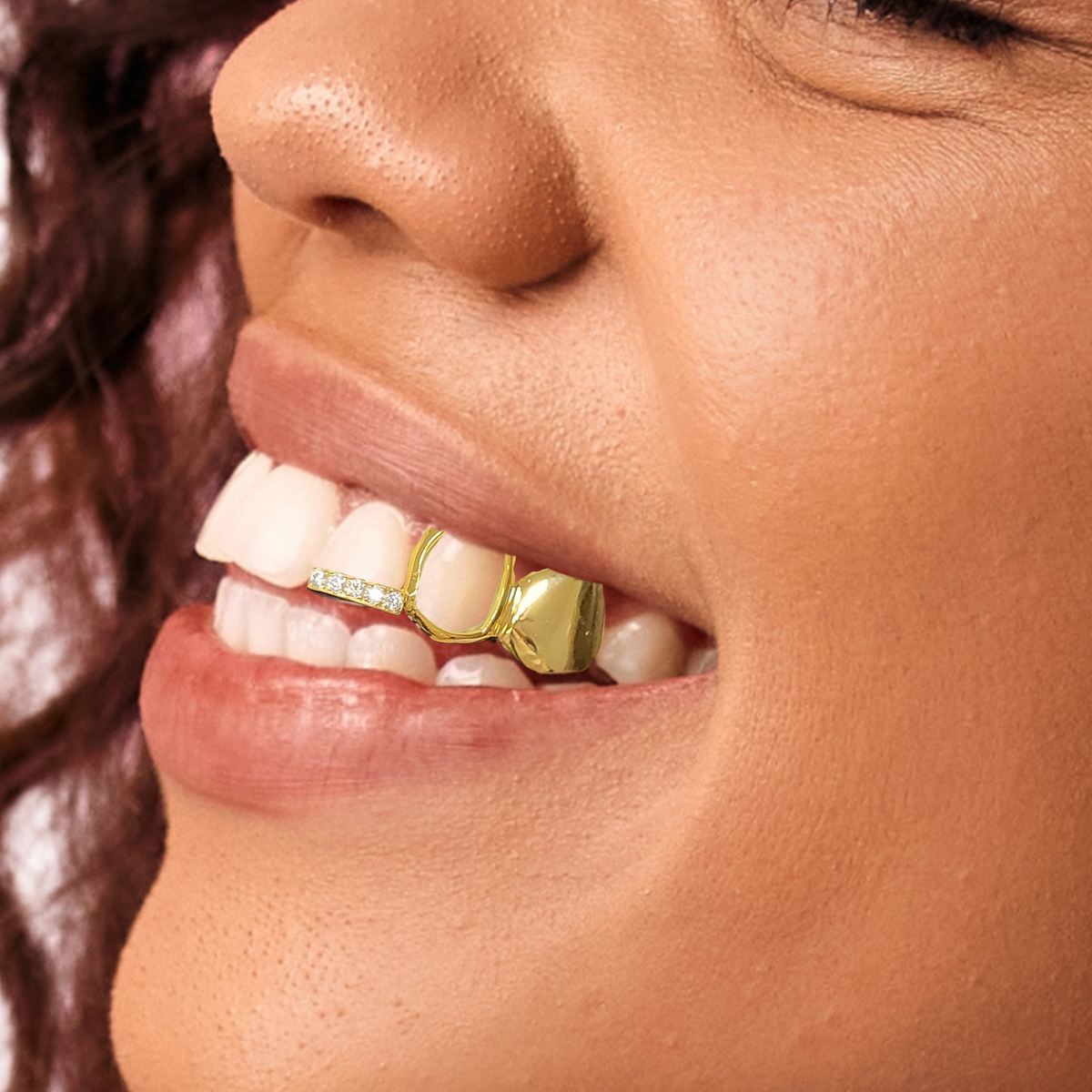 A close-up of a person's mouth wearing gold teeth grills on the front teeth, with one tooth featuring a large open face and another lined with small clear stones.