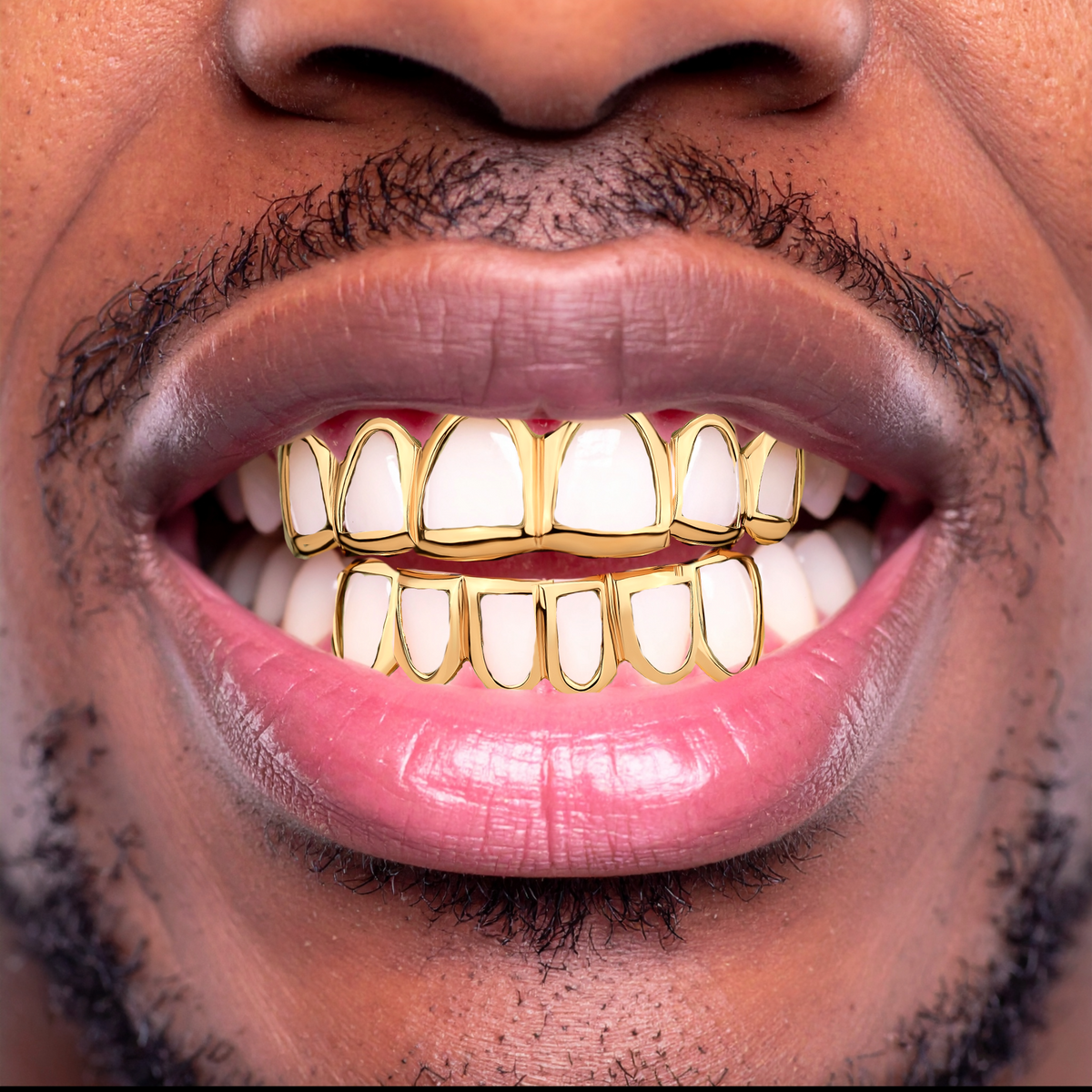 A close-up of a smiling man's mouth wearing gold grillz on his teeth, with visible skin, lips, and facial hair.