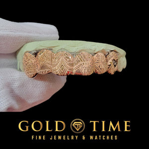 Rose gold 6 piece grillz with Polynesian tribal engraving held on a dental model in a gloved hand
