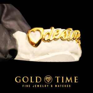 Gold nameplate grill with an open heart canine and cursive name, held on a dental mold