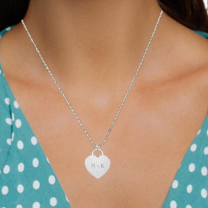 White gold heart disc charm engraved with initials, worn on a chain by a person in a teal polka dot dress