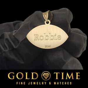 Satin back of a gold football pendant engraved with the name Robbie, held in a black gloved hand